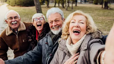 Fyra glada äldre vuxna skrattar och ler medan de tar en selfie utomhus i en park.