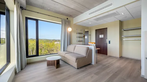 A bright, modern room with a large window offering a scenic view of lush greenery. A comfortable sofa and a small table furnish the space.