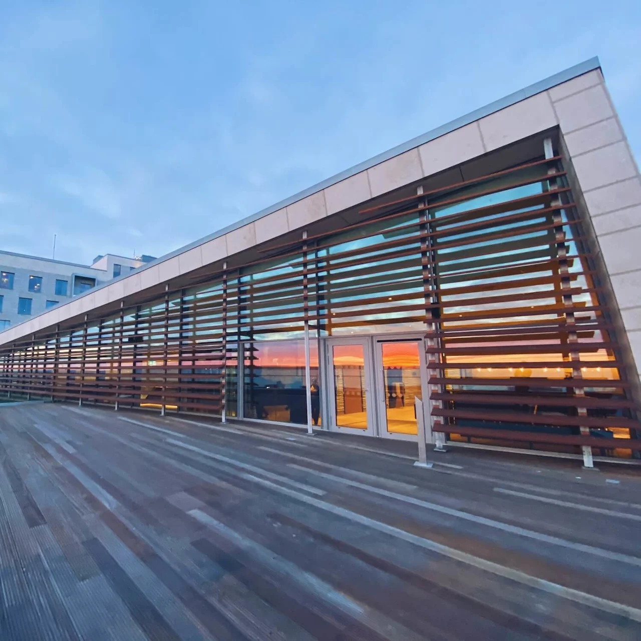 En moderne bygning med store trespalter og glassvinduer reflekterer en fargerik solnedgang, sett fra en romslig utendørs trebalkong, under en klar himmel.