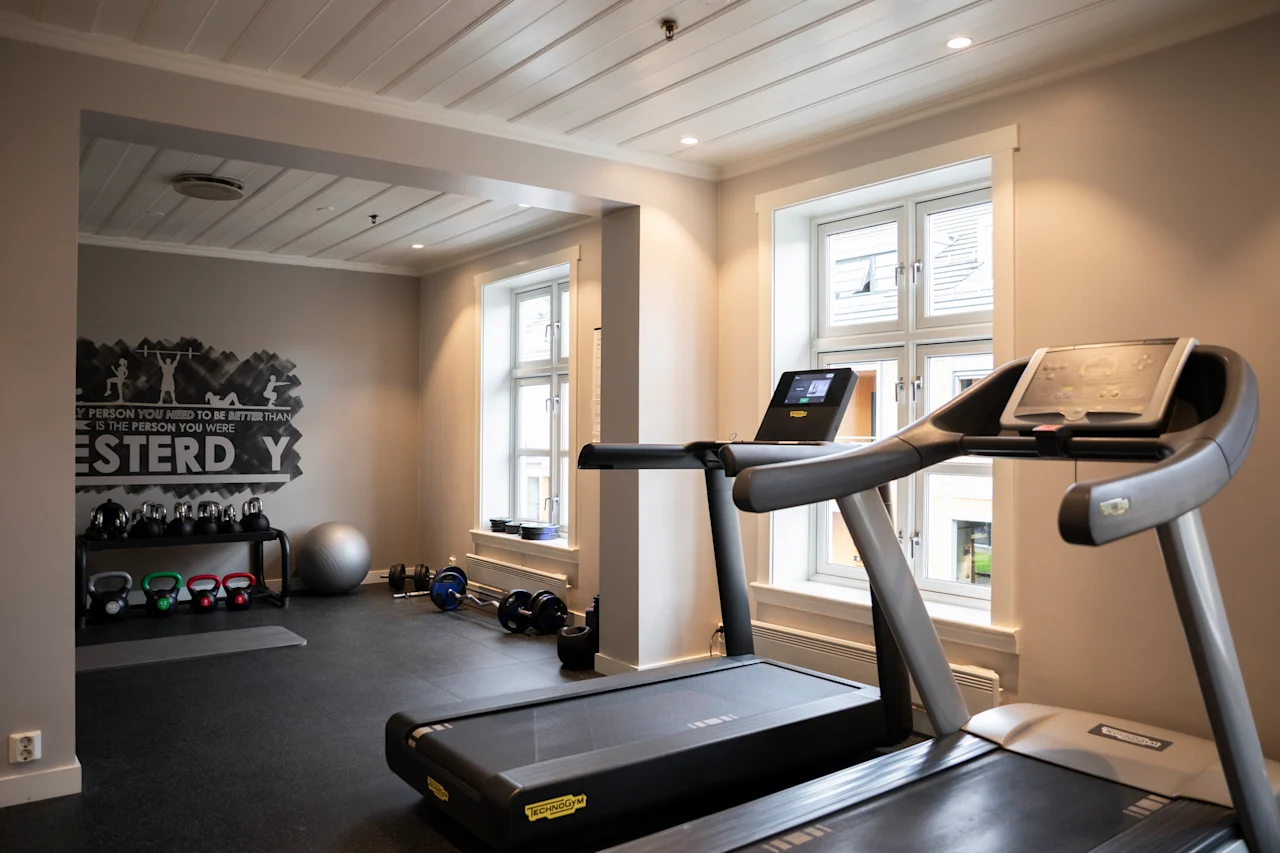 Løbebånd står ubrugt i et lyst fitnessrum med lyse vægge. Bag det hviler kettlebells og en træningsbold på gulvet tæt på en motiverende vægmaleri: "EVERY PERSON YOU KNOW USED TO BE A STRANGER. THE PERSON YOU ARE IS UP TO YOU."