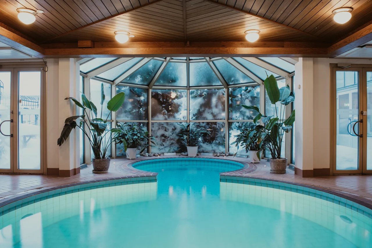 Ett inbjudande inomhuspoolområde med bubbelpool, omgivet av frodiga gröna växter och stora fönster, under ett varmt trätak.