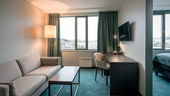 En beige sofa står i et godt oplyst rum ved siden af en høj lampe og et træ-sofabord, med udsigt udenfor. Et skrivebord med stol og TV er i nærheden.