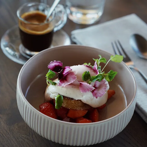 En vackert upplagd efterrätt med jordgubbar, grädde och ätbara blommor, serverad med en kopp kaffe och vatten.
