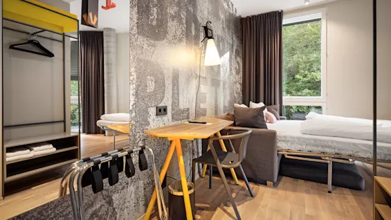 A small, modern room features a desk with a lamp against a textured wall, a bed near a large window with curtains, and a minimal wardrobe. 