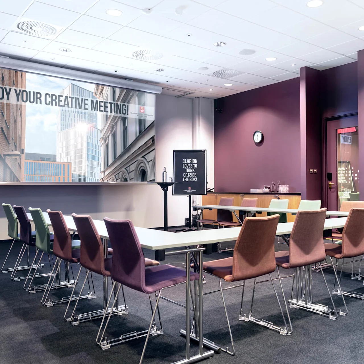 Et konferanserom med fargerike stoler rundt bord; en stor skjerm viser "ENJOY YOUR CREATIVE MEETING!" og et skilt lyder "CLARION LOVES TO THINK OUTSIDE THE BOX" mot lilla vegger.