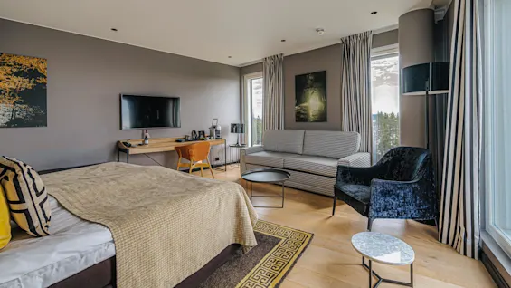 A neatly arranged hotel room features a bed with patterned pillows and blanket, a TV on the wall, a desk with a chair, and a sofa beside large windows.