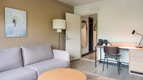 Gray sofa faces round wooden coffee table; standing lamp beside it. Open door reveals hallway. Desk with lamp, chair, and coffee station placed against the right wall. Artwork labeled "BIØVIK" on left.