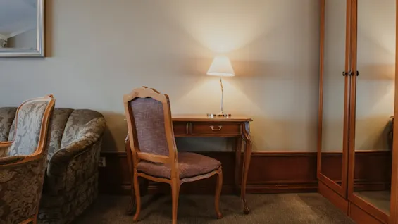 Ett mysigt rum med klassiska möbler, inklusive ett träskrivbord, stol och en mönstrad soffa, upplyst av en varm lampa.