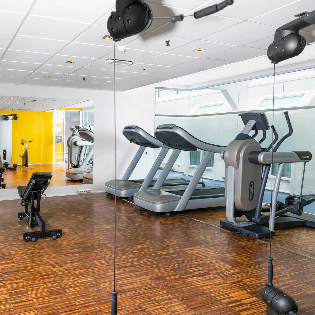 A gym with treadmills, a stationary bike, and weights. A bench faces a mirrored wall reflecting yellow accents. Brown wooden flooring and white walls create a clean ambiance.