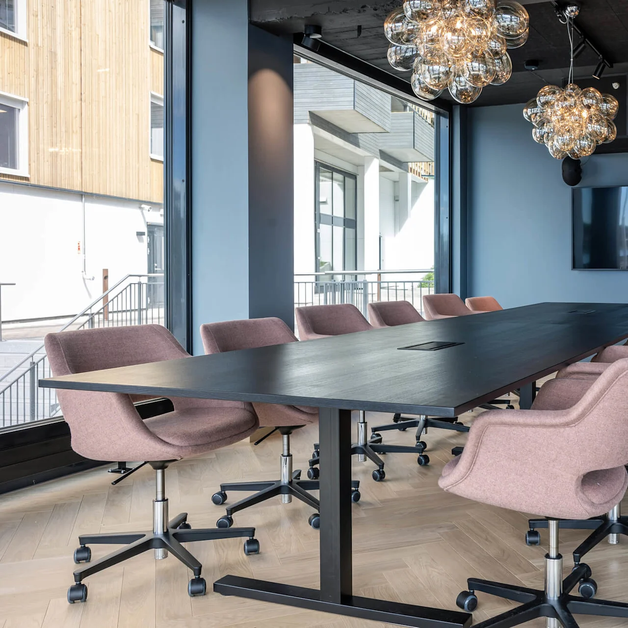 Et langt sort konferencenbord er omgivet af lyserøde kontorstole med hjul i et moderne mødelokale med store vinduer og hængende lysarmaturer.