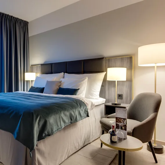 En stor seng med teal sengetøy står i et moderne, godt opplyst hotellrom. Et vindu avslører en naturskjønn utsikt over en marina og fjell. Et sidebord har en meny og en liten vinflaske.