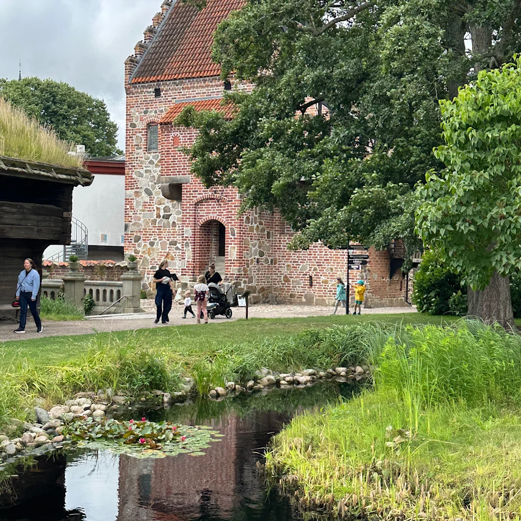 People stroll past a historic brick building with a red roof, reflected in a tranquil pond with water lilies, surrounded by lush greenery.