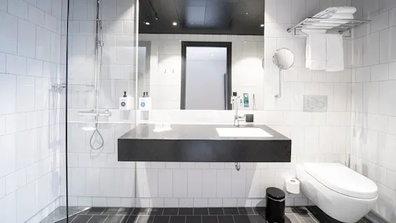 Bathroom with white tiled walls features a glass shower, black countertop sink, and wall-mounted toilet. Towels hang on a rack above. Two dispensers labeled "I am a soap" and "I am a shampoo."