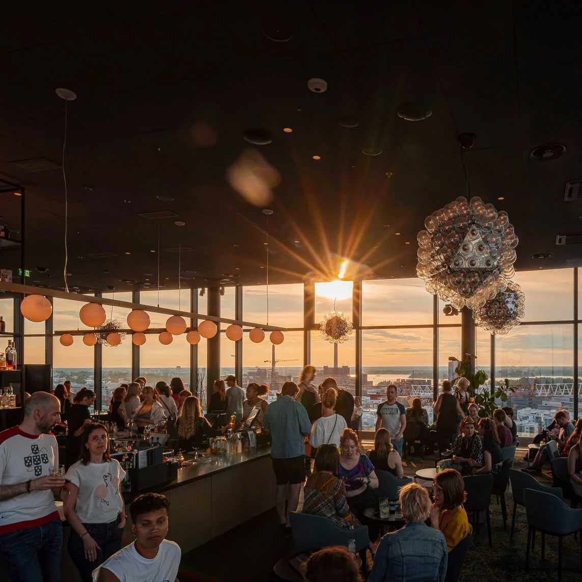 Bargæster nyder solnedgangsudsigter gennem store vinduer, med glødende sfæriske lys over hovedet. Helsingfors skyline er synlig, og "HELSINKI" er oplyst på en hylde.