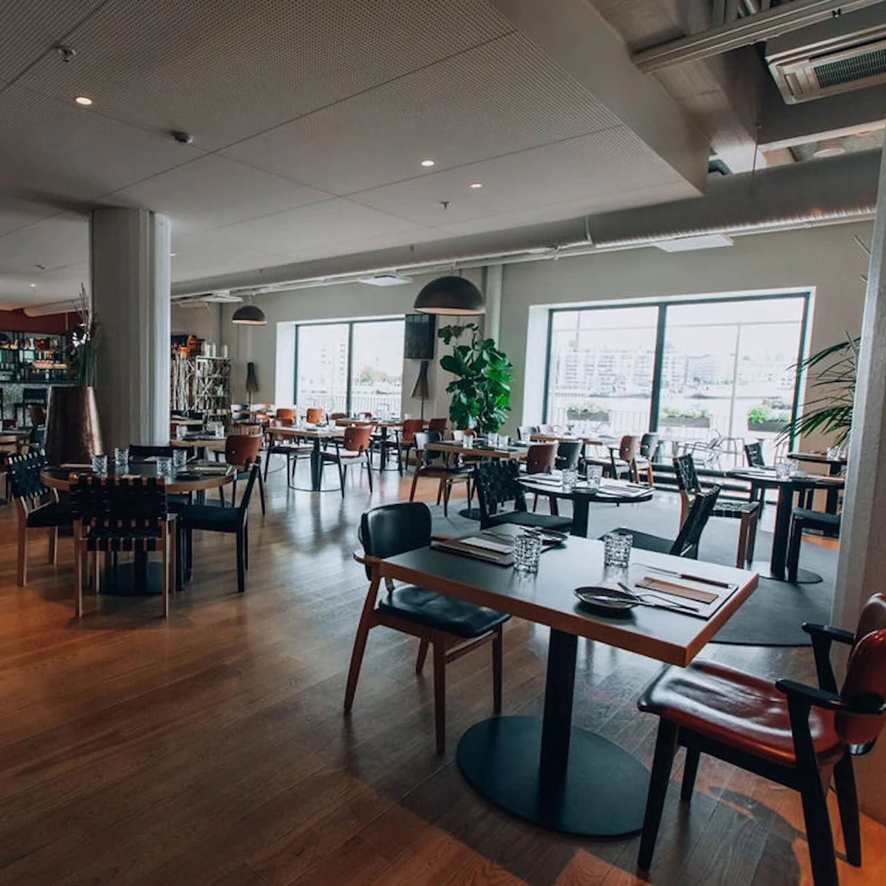 Restaurantbord pent arrangert, med minimalistisk dekor og tregulv. Store vinduer gir naturlig lys med utsikt over utendørsområdet. Atmosfæren er romslig og moderne, ideell for bespisning.