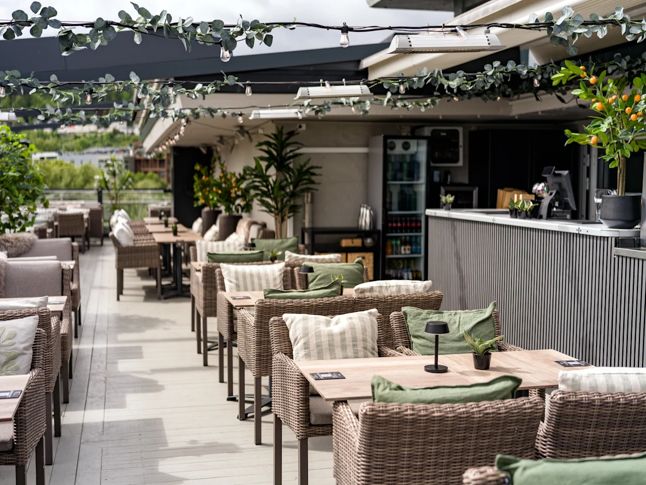 Cafébord med korgstolar är placerade på en takterrass. Ljusslingor och växter skapar en mysig stämning i en modern miljö med utsikt över ett avlägset landskap.