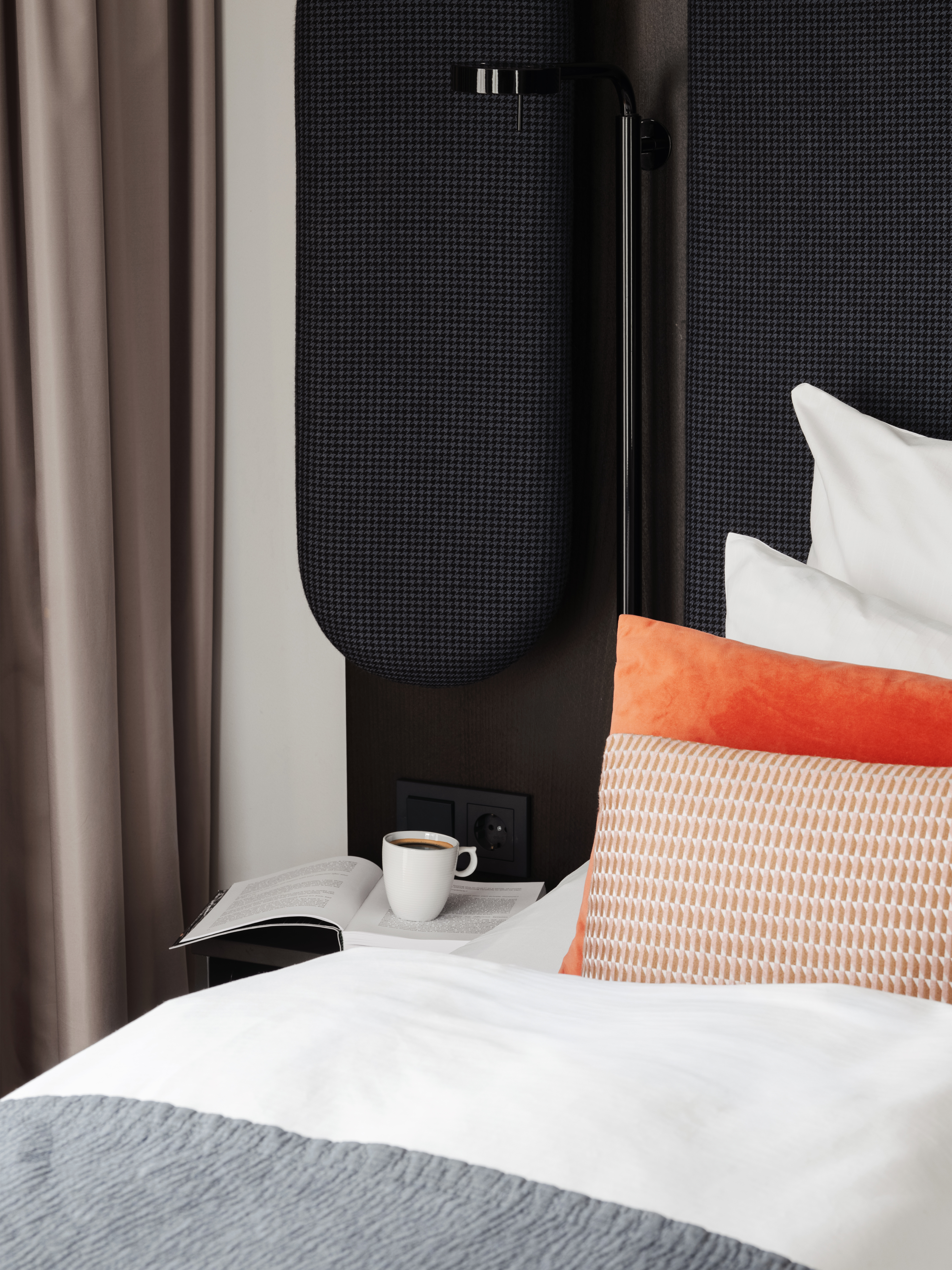 En hyggelig scene ved sengen med en åben bog og en kop kaffe på et natbord ved siden af en seng med puder.