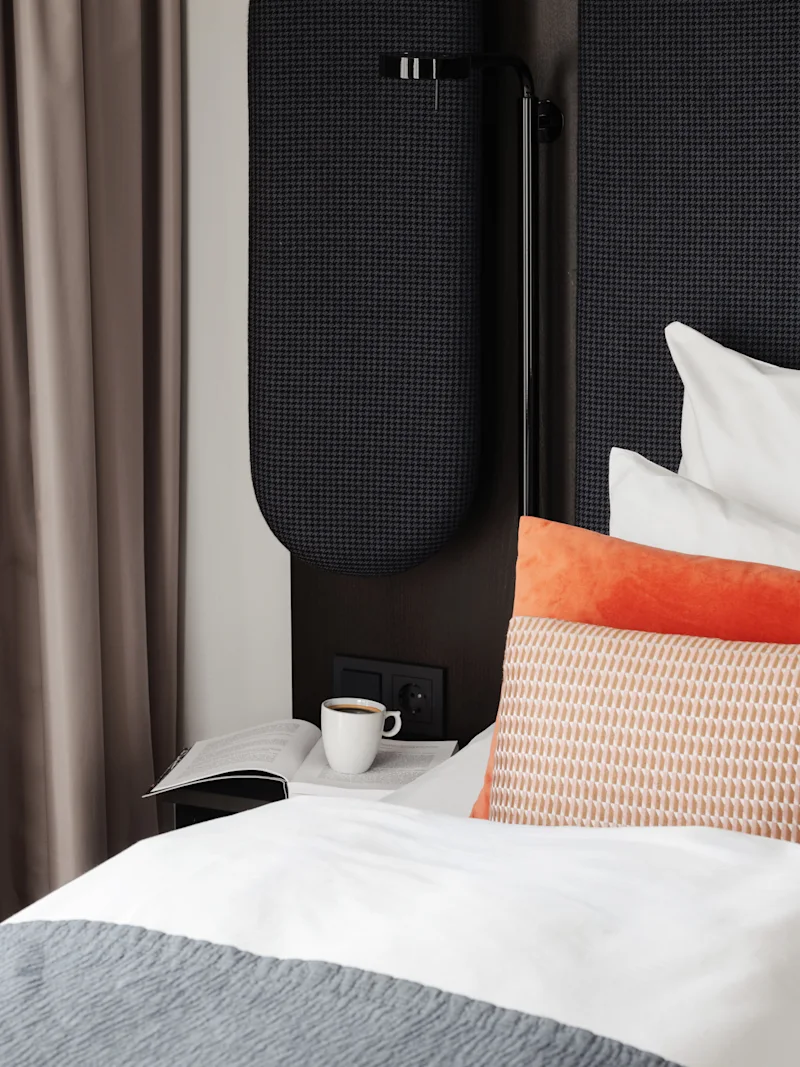 En hyggelig scene ved sengen med en åben bog og en kop kaffe på et natbord ved siden af en seng med puder.