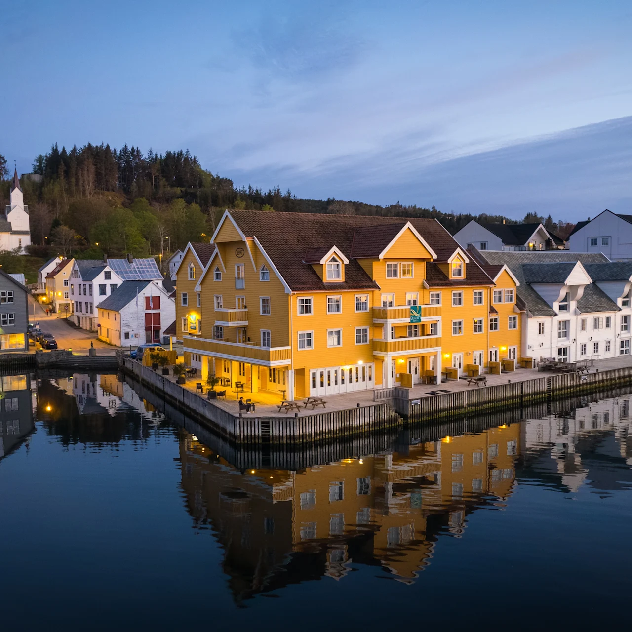 Colorful buildings line a calm waterfront, their reflections shimmering in the water under a twilight sky. Boats are docked nearby, and a forested hill rises in the background.
