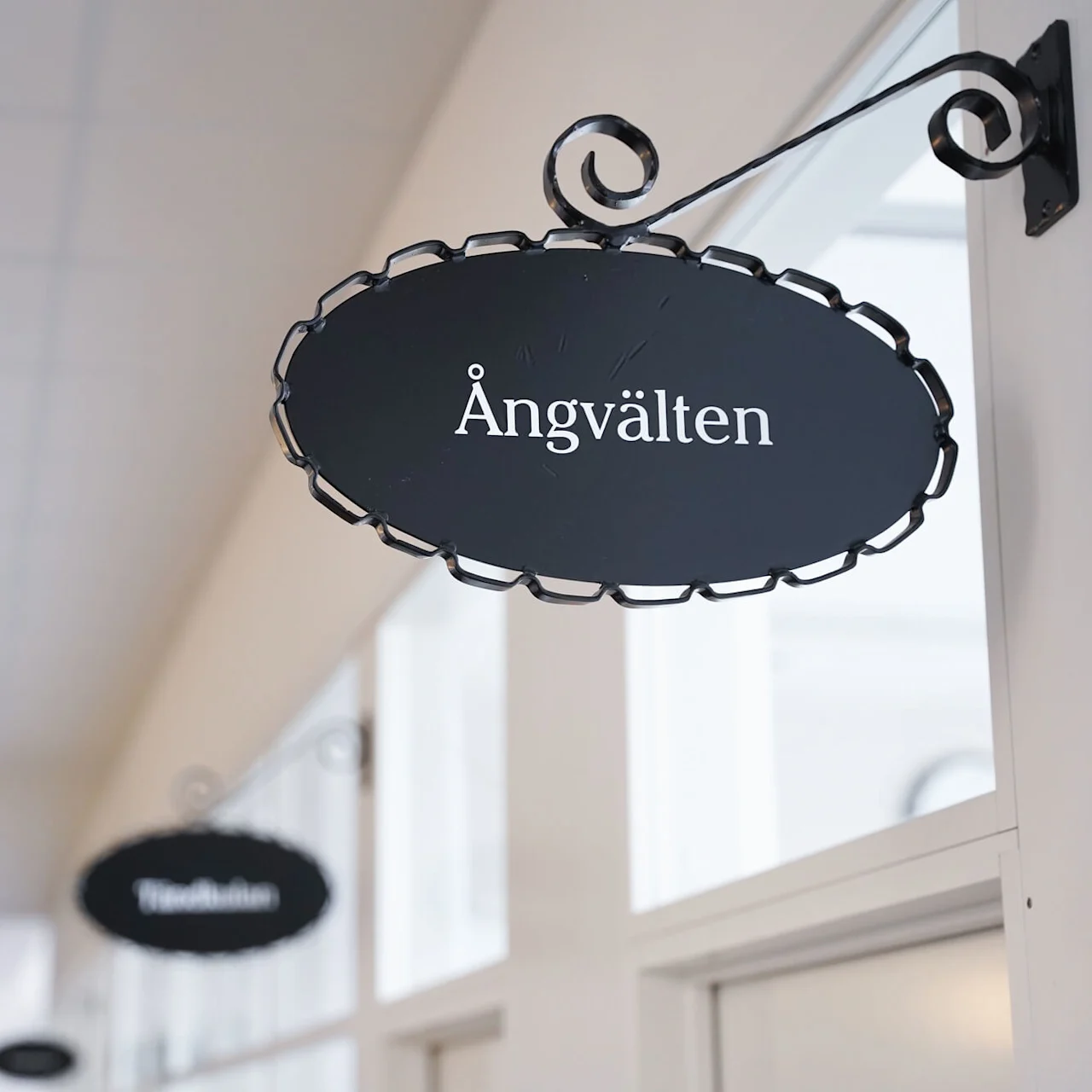 En oval skylt hänger på en metallkonsol med texten "Ängvälten" i en inomhusmiljö.