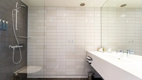 Et moderne badeværelse har en bruseniche med grå fliser til venstre og en dobbelt hvid vask med spejl til højre. Håndklæder og toiletartikler er pænt arrangeret.