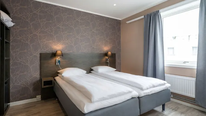 A double bed with white linens is neatly arranged against a patterned wallpaper, under soft lighting. A window with gray curtains provides natural light, set in a modern bedroom.