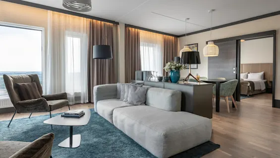 A gray sofa sits on a blue rug alongside a small coffee table, in a modern living room with large windows, soft curtains, and stylish lamps. Adjacent is a dining area leading to a bedroom.