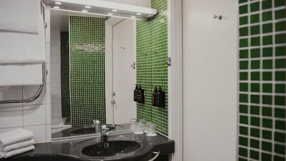 Siisti ja moderni kylpyhuone Home Hotel Uppsalassa vihreillä mosaiikkilaatoilla, pesualtaalla, peilillä ja puhtailla pyyhkeillä.