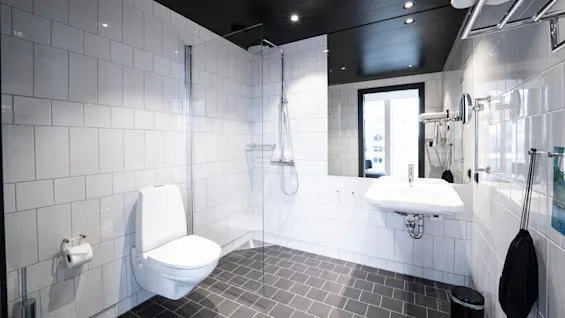 A modern bathroom with a white toilet and sink, a glass-walled shower area, and black tiled floor. Towels hang on a rack near the ceiling, under recessed lighting.