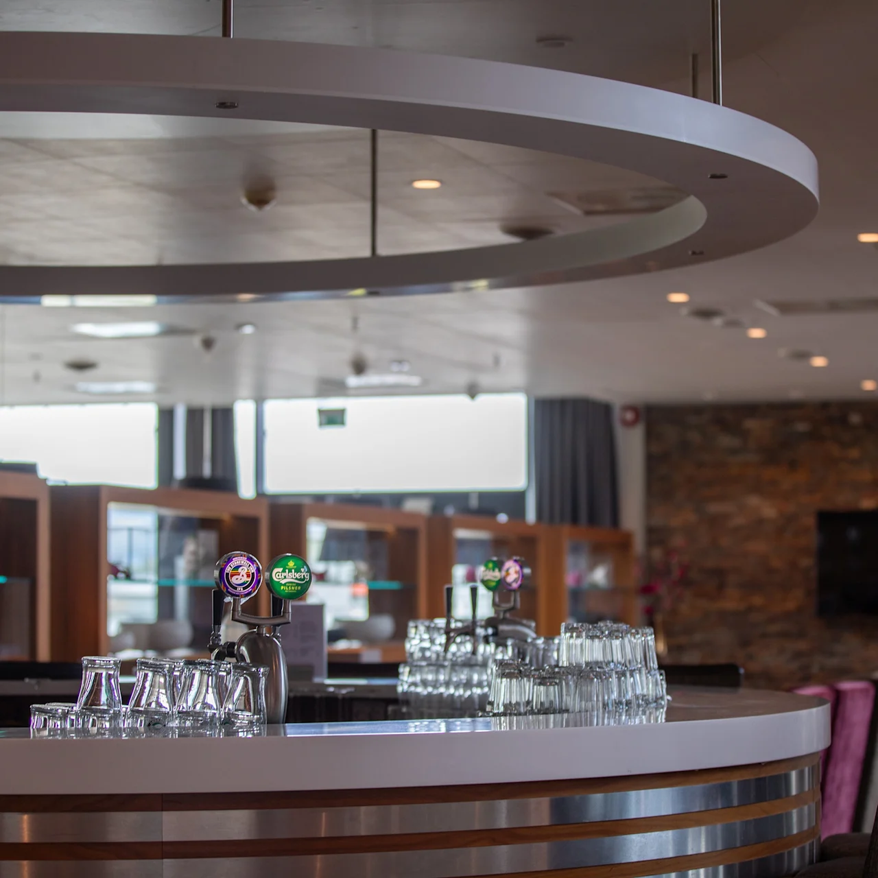 Bar med tappekraner og glass på en glatt benk; omgitt av stoler og stilige dekorasjoner i et moderne, godt opplyst salongområde. Ingen tekst til stede.