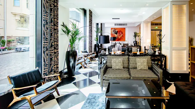 En elegant salong med mønstrede sofaer og et glasbord i moderne omgivelser med store planter, oplyst af naturligt lys fra gulv-til-loft-vinduer.