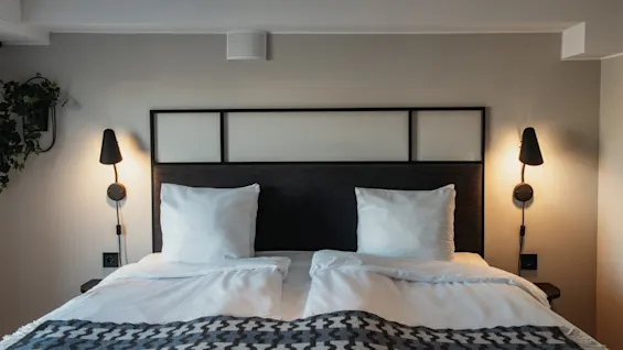 Et hyggeligt soveværelse med en pænt redt seng, bløde puder og varm belysning fra væglamper, der indbyder til afslapning.