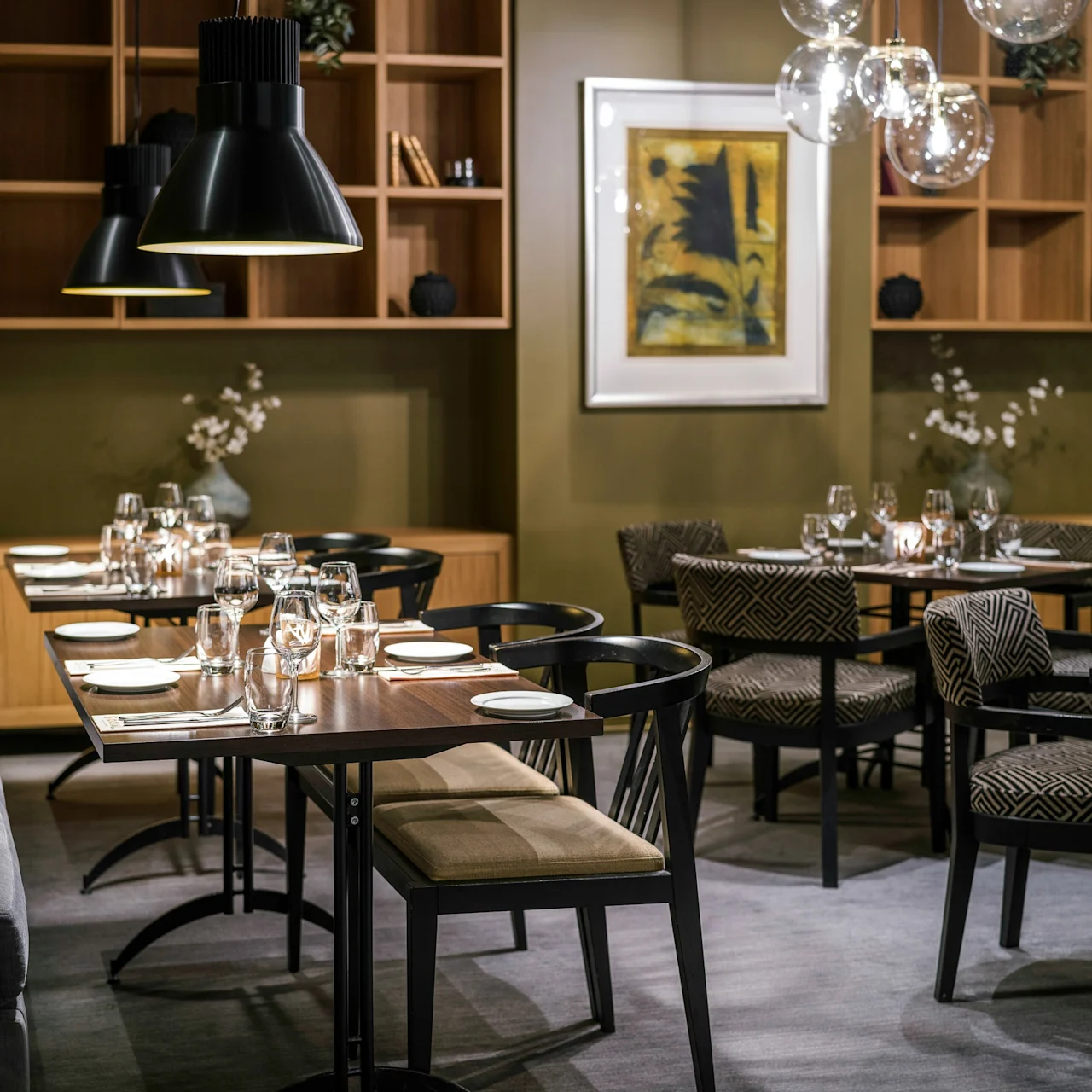 Tables are set with glassware and plates in a modern restaurant, surrounded by stylish chairs and soft lighting from overhead lamps, creating an elegant dining atmosphere.