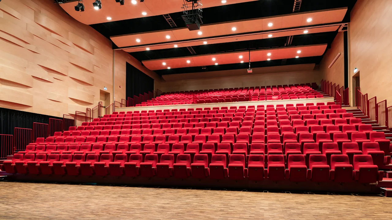 En stor, tom sal med rader av livlige røde seter vendt mot en trescene. Veggene er i lyst tre med teksturerte paneler.
