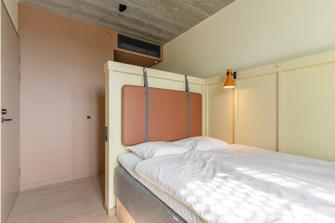 Säng i ett litet rum, täckt med vita lakan. En orange vägglampa är monterad ovanför sänggaveln. Ljus strömmar in genom ett osynligt fönster och kastar skuggor.