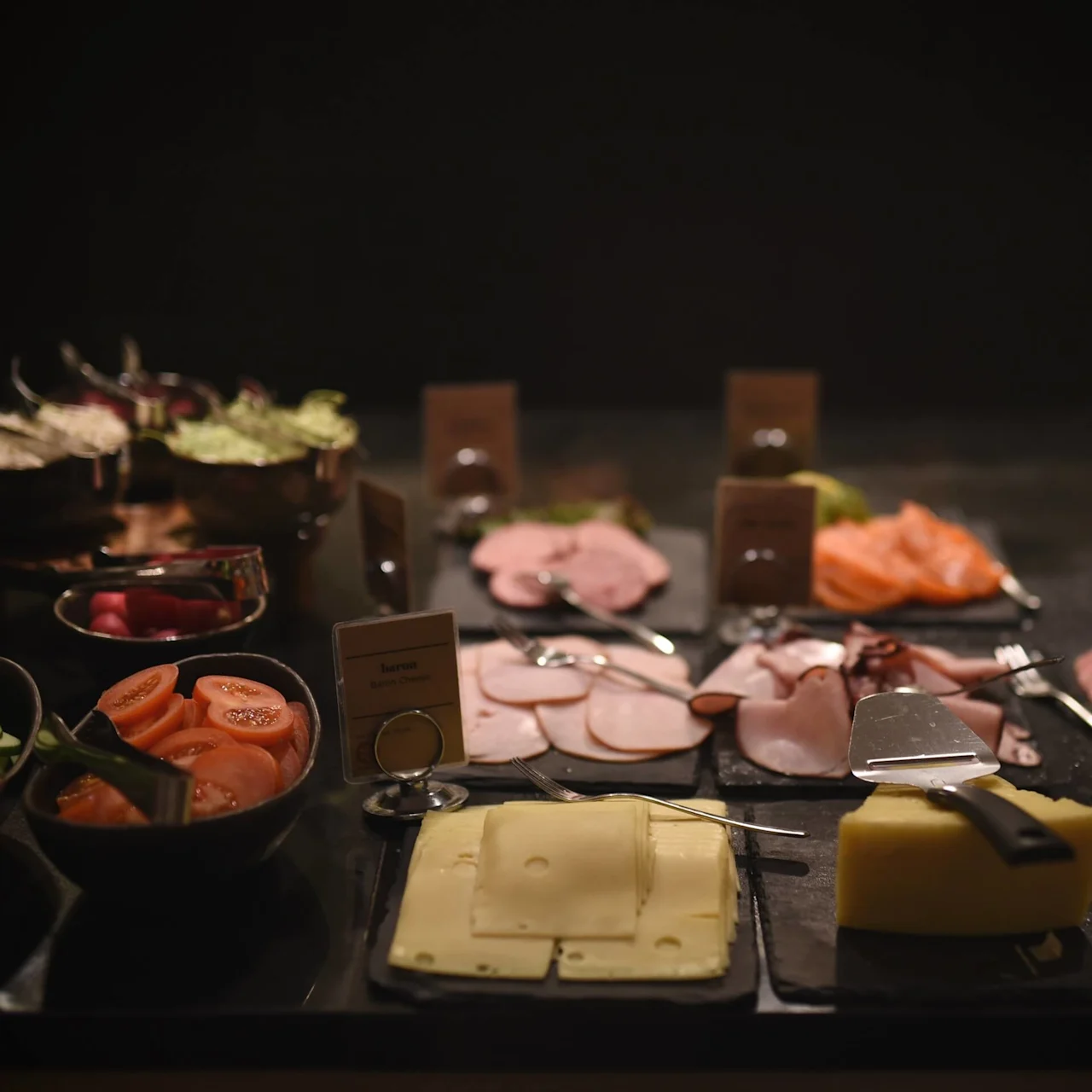 En buffébord håller skivade grönsaker, ostar och kött, alla elegant arrangerade. Kort etiketterar föremålen, inklusive "baron Gourmet Cheese" och "brie ost Sur La Route."