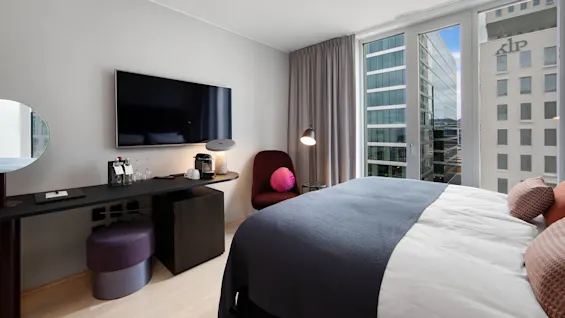 TV mounted on a wall, above a desk with drinks, glasses, and a coffee machine; adjacent to a stylish chair with a pink cushion. Large window reveals city buildings.