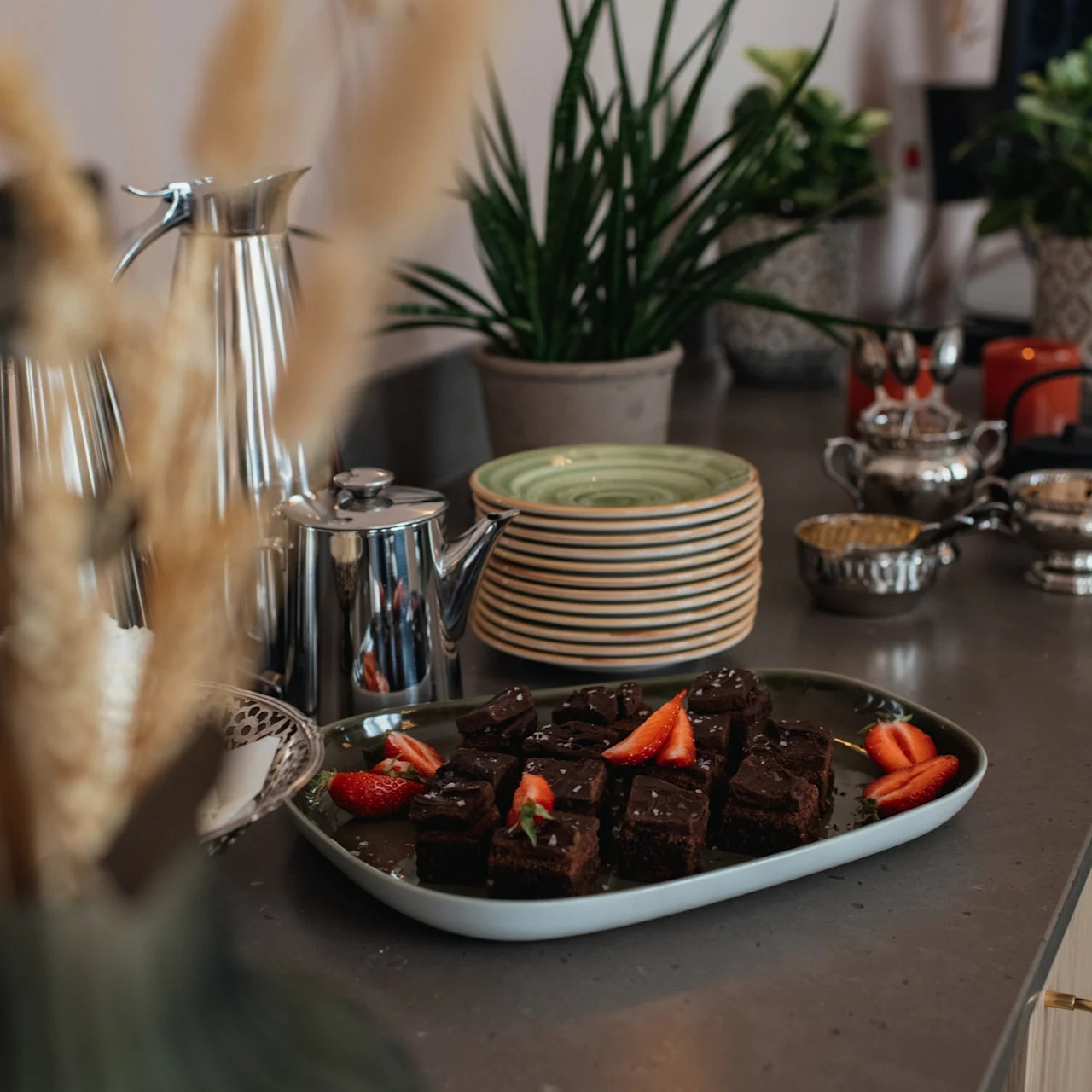 A tray of chocolate brownies topped with sliced strawberries rests on a kitchen counter. Surrounding are plates, silver coffee pots, and potted plants creating a cozy kitchen setting.
