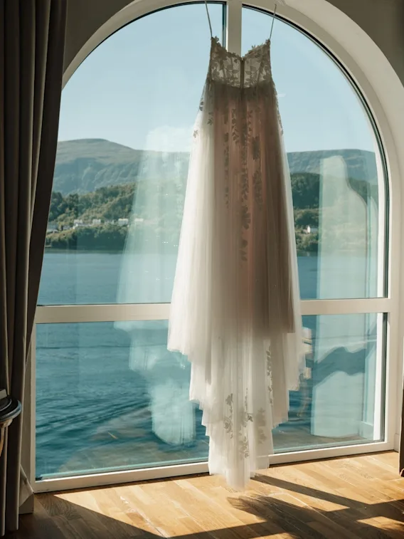 En hvit brudekjole i blonder henger foran et stort buet vindu med utsikt over fjord og fjell i Ålesund.