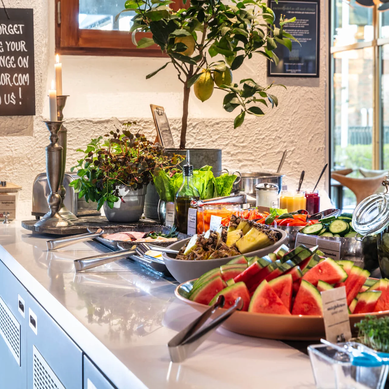 A buffet table spread with fresh fruits, meats, and salad in a well-lit room. A sign encourages sharing on TripAdvisor.