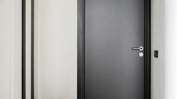 En smal gang med en minimalistisk stil har en svart dør, et høyt speil og en rund, svart krakk på tregulv. Myk belysning tilfører varme til det enkle, moderne designet.
