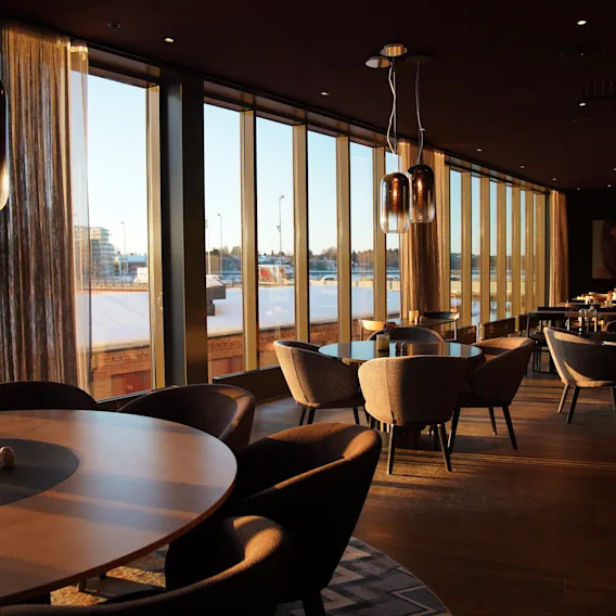 Moderne restaurant eller lounge med store vinduer mod snedækket landskab ved solnedgang, med stilfulde borde og stole.