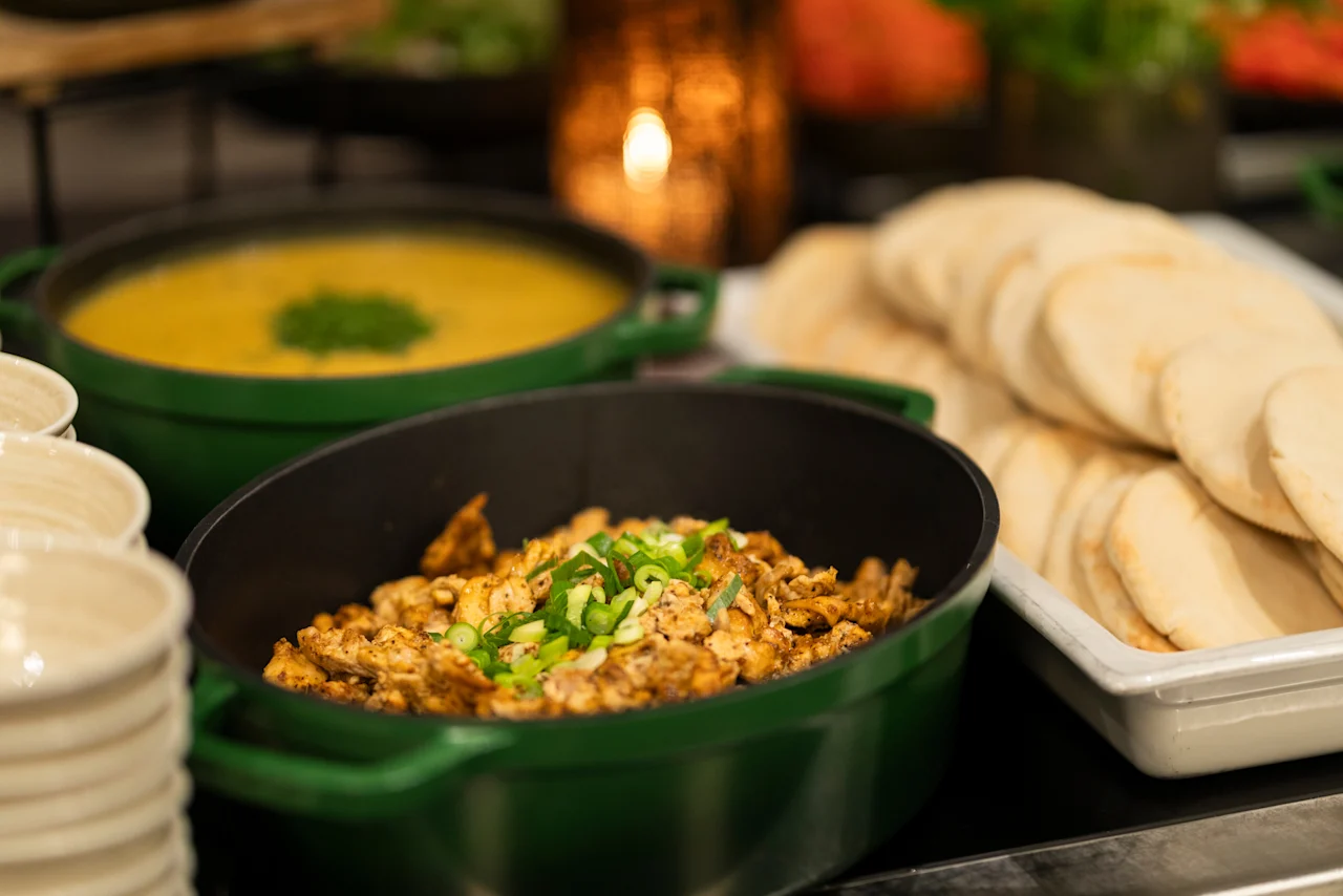 A buffet spread at Home Hotel Grand Bodø featuring a yellow soup, seasoned chicken, and flatbreads, with stacked bowls ready for guests.
