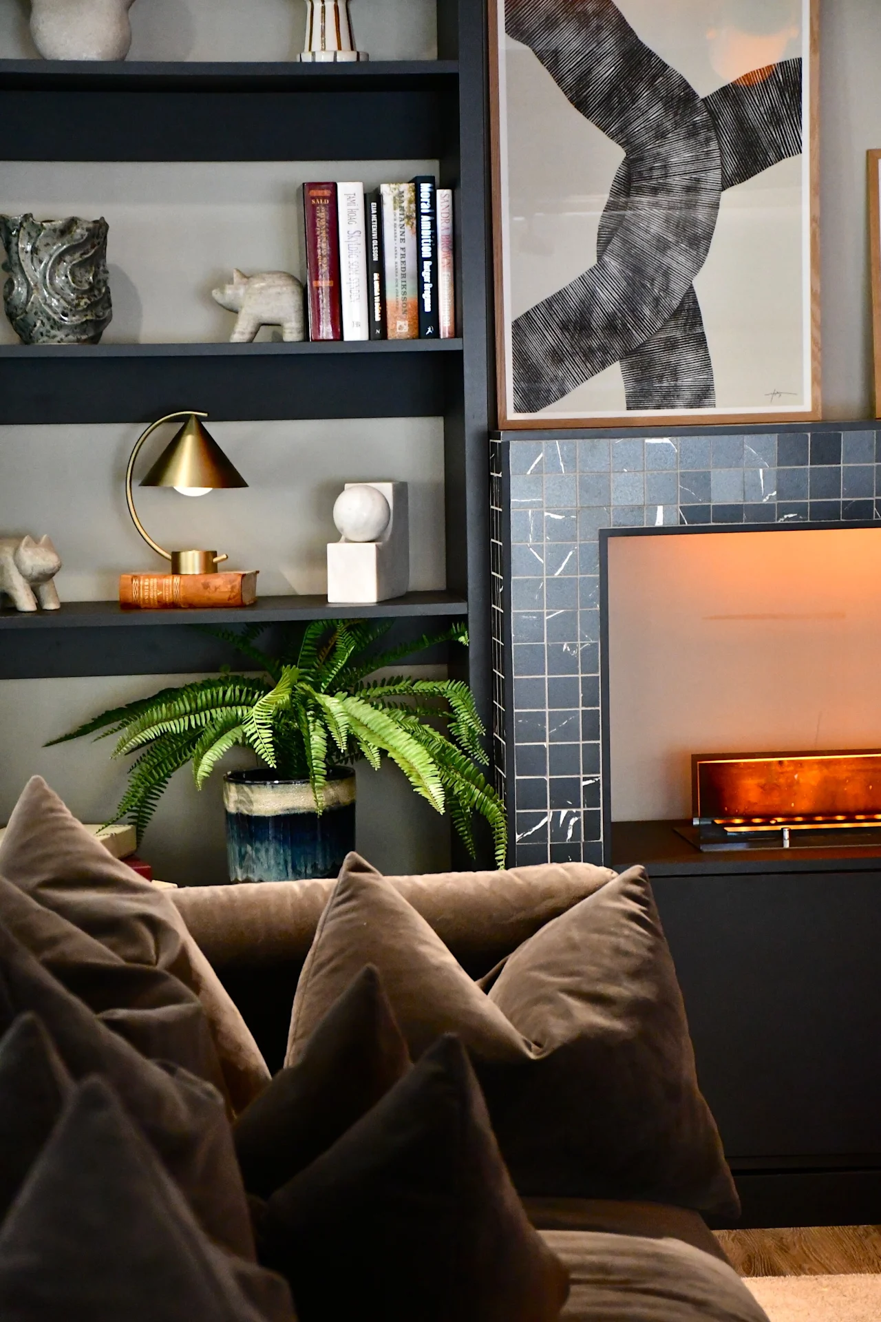 Et varmt og indbydende opholdsrum med en plyssofa, en bogreol med bøger, indretning, en plante og en glødende pejs.