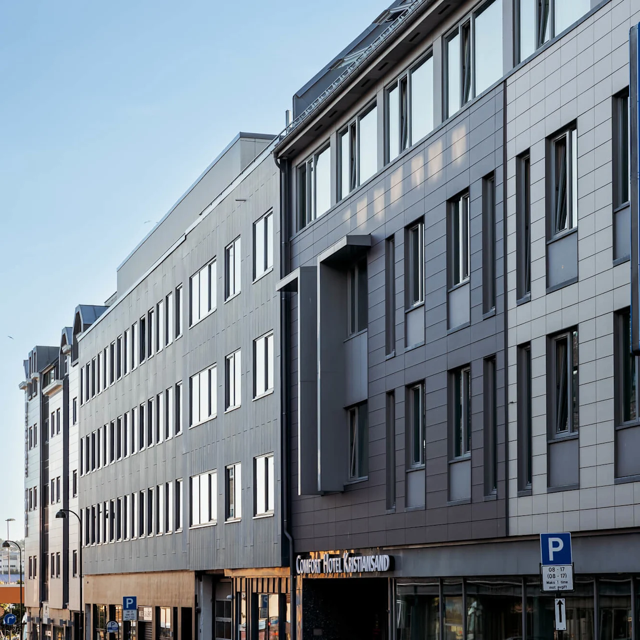 A multi-story hotel building with a blue sign reading "COMFORT HOTEL" and "Comfort Hotel Kristiansand." It is situated on a street with parked cars. Parking sign states "08–17 Man–lør 1t max privat."  