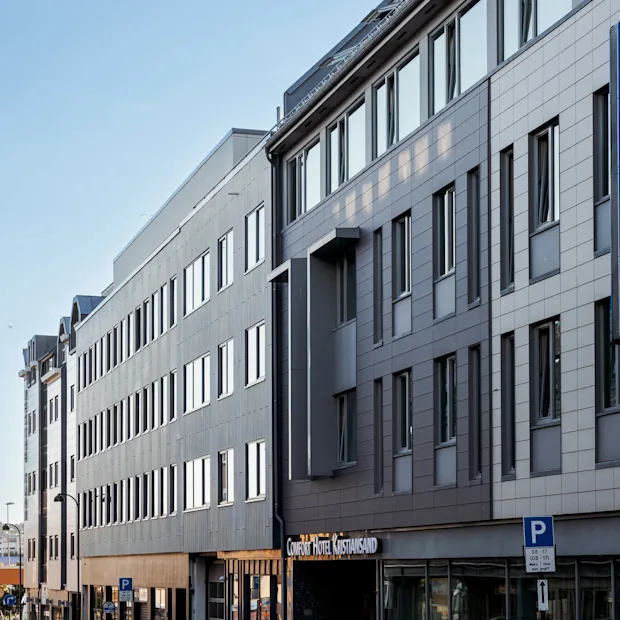 En fleretasjes hotellbygning med et blått skilt med teksten "COMFORT HOTEL" og "Comfort Hotel Kristiansand." Det ligger på en gate med parkerte biler. Parkering skiltet sier "08–17 Man–lør 1t max privat."