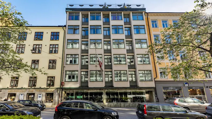 A modern building with large windows and a rooftop terrace bathed in sunlight, situated in an urban street with cars parked in front.