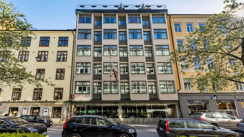 A modern building with large windows and a rooftop terrace bathed in sunlight, situated in an urban street with cars parked in front.
