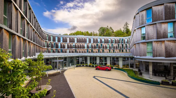 Modern hotel facade with curved wooden panels surrounds a courtyard, featuring a parked red car. Trees and plants add greenery. Signs read "CLARION" and "RESTAURANT."