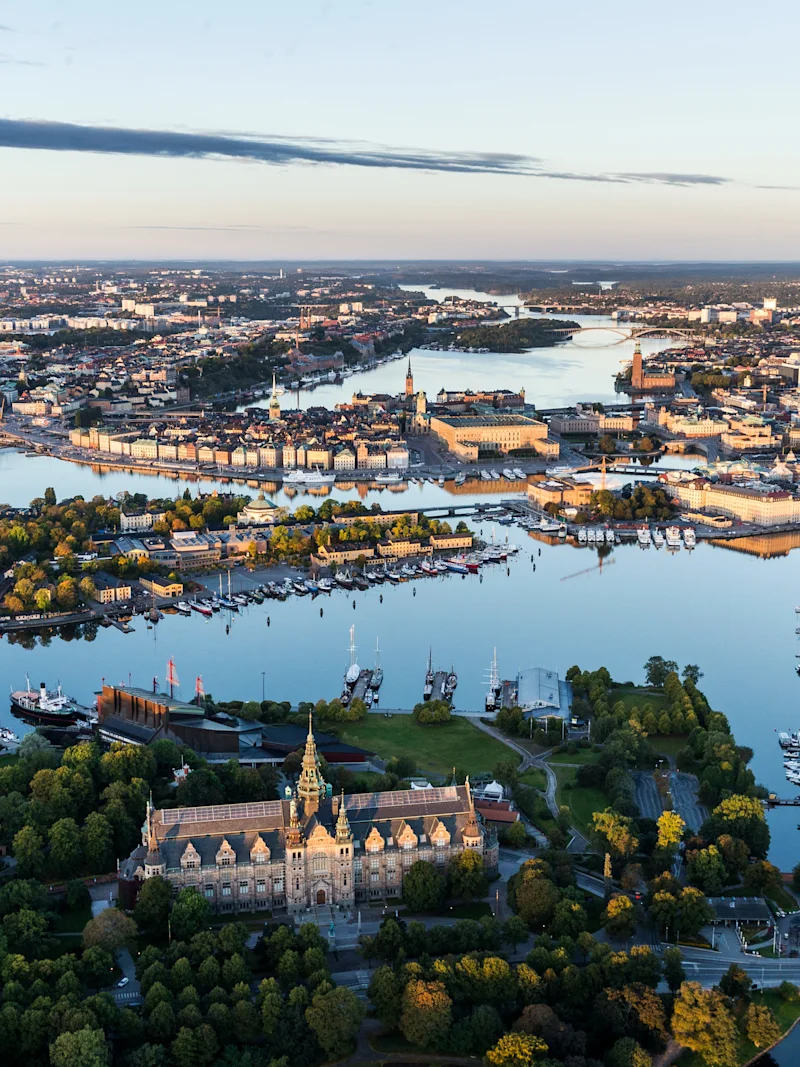 A historic building is surrounded by lush trees and water, nestled within an urban landscape. Numerous boats are docked along the waterfront, under a vast, clear sky.